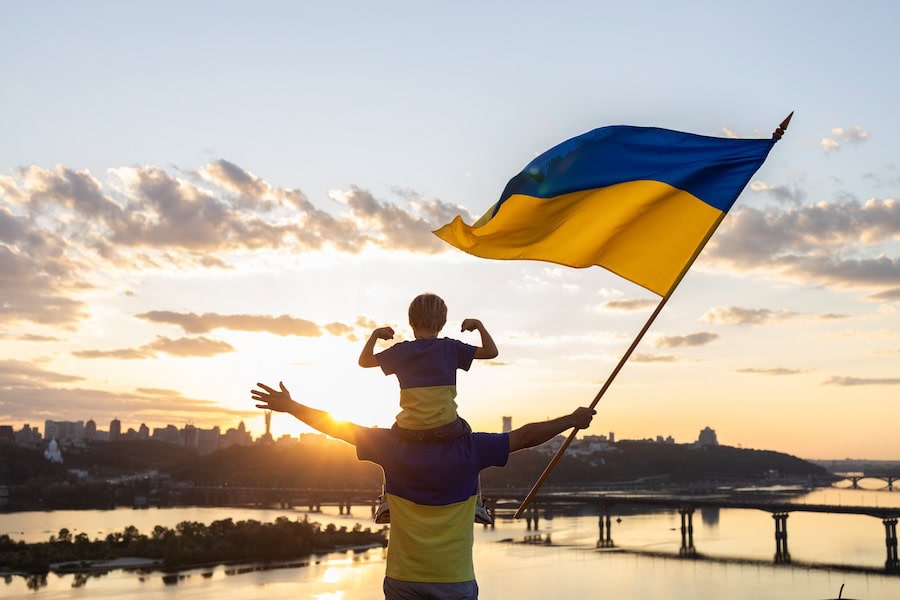 dad with son, who sits on shoulders, with large flag of Ukraine on roof of house on embankment of Dnieper River